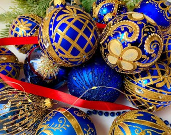 May include: A collection of blue and gold Christmas ornaments. The ornaments have intricate gold designs, including floral patterns and geometric shapes. A red ribbon and greenery are also present, creating a festive holiday scene.