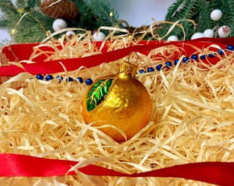 May include: A gold Christmas ornament with a green and gold leaf design, resting on a bed of straw. Red ribbon and blue beads are also visible, with a backdrop of pine branches and lights. Festive holiday decor.