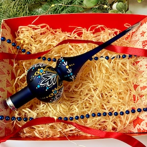 Op de afbeelding: Een donkerblauwe kerstboomtop met witte, gele en blauwe bloemmotieven. De top zit in een rood-wit patroon doos met stro, rood lint en blauwe kralen slinger. Een feestelijke vakantiedecoratie.