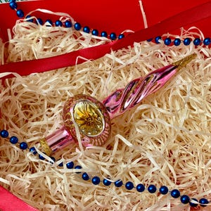 May include: A pink and gold Christmas tree ornament with a spiral design, resting on shredded paper inside a red box. Blue beaded garland and a red ribbon add festive accents. The ornament has a gold glittered tip.
