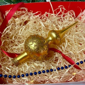 Op de afbeelding: Een gouden kerstboomtop met een decoratief ontwerp, rustend in een rood-wit patroon doos gevuld met stro. Een rood lint en een blauwe kralenketting liggen ook in de doos.