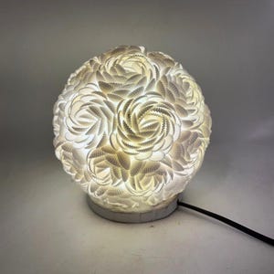 May include: A white, spherical lamp with a textured surface, resembling swirling petals. The lamp emits a soft, warm glow, and has a circular base with a black power cord. The design is intricate and decorative, suitable for a living space.