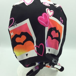May include: Black scrub hat with a colorful print of hearts and instant photo frames. The hat has a tie at the back.