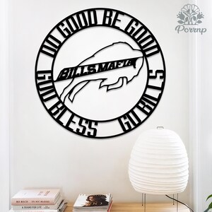 May include: A black metal wall art piece featuring the words "DO GOOD BE GOOD" and "GO BILLS" encircling a buffalo image with "BILLS MAFIA" across it. The artwork is mounted on a white wall, with a white lamp and books below.
