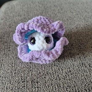 Puede incluir: Una almeja de ganchillo hecha a mano con un interior blanco y afelpado y una concha lavanda. El interior de la almeja es de color azul brillante. La almeja tiene ojos saltones grandes y negros.