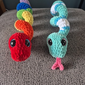 Puede incluir: Dos serpientes de crochet de peluche. Una es roja con un cuerpo arcoíris, la otra es turquesa con remolinos blancos y azules. Ambas tienen ojos grandes y negros y lenguas rosas. Las serpientes están sobre una superficie gris texturizada.