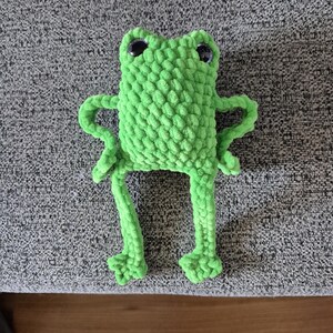 Puede incluir: Una rana de ganchillo verde brillante hecha a mano con grandes ojos negros. La rana tiene los brazos extendidos y patas largas con pies detallados. El peluche está hecho de un hilo suave y texturizado y se encuentra sobre una superficie gris.