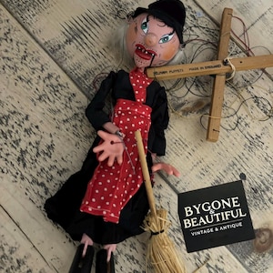 Könnte beinhalten: Eine Vintage-Pelham-Marionette einer Hexe, gekleidet in einem schwarzen Kleid und einer roten gepunkteten Schürze. Die Marionette hat ein bemaltes Gesicht, einen schwarzen Hut und einen Strohbesen. Die Holzsteuerung ist mit "MADE IN ENGLAND" gekennzeichnet.