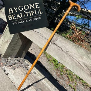 May include: A brown wooden walking stick with a curved handle. The stick is resting on a wooden fence. The text "Bygone & Beautiful Vintage & Antique" is visible in the background.