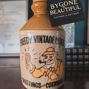 Può includere: Una bottiglia di ceramica marrone con un tappo di sughero e un'etichetta che recita "Creedy Vintage Cider Greetings from Cornwall". L'etichetta presenta un'illustrazione a fumetti di un uomo con una pipa e un bicchiere di sidro.
