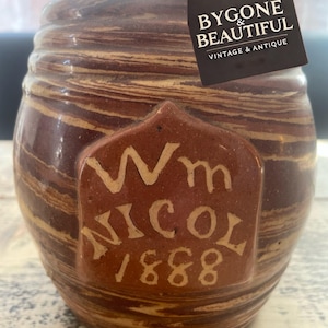 Puede incluir: Un tarro de gres vintage marrón y beige, de forma cilíndrica. Presenta un emblema rectangular con el nombre "Wm NICOL 1888" en un tono más claro. Lleva una etiqueta que dice "BYGONE & BEAUTIFUL VINTAGE & ANTIQUE".