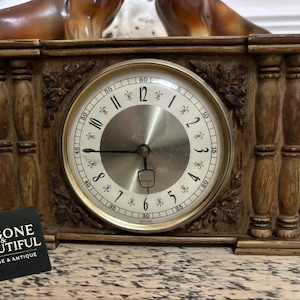 May include: A vintage wooden mantel clock with a round face and gold-tone trim. The clock has a light-coloured face with black numerals and hands. The clock is decorated with carved details and the text "BYGONE & BEAUTIFUL VINTAGE & ANTIQUE".