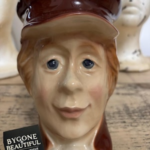May include: A vintage ceramic mug shaped like a smiling person wearing a brown cap. The mug has a tan face with rosy cheeks and blue eyes. The logo "BYGONE & BEAUTIFUL VINTAGE & ANTIQUE" is visible on a black tag.
