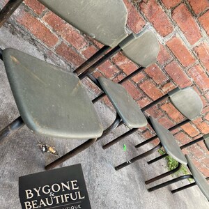 May include: A row of vintage folding chairs with olive green seats and black metal frames. The chairs are arranged on a concrete surface with a brick wall in the background. A sign that reads "BYGONE & BEAUTIFUL VINTAGE & ANTIQUE" is in the foreground.