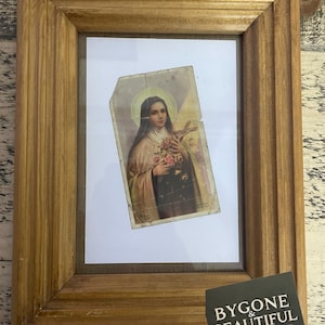 Könnte beinhalten: Ein gerahmter Vintage-Religionsdruck mit einer Frau, die ein Kreuz und Blumen hält. Der Druck ist in einem hellbraunen Holzrahmen mit gerilltem Design eingefasst. Ein Aufkleber in der unteren rechten Ecke lautet "BYGONE & BEAUTIFUL VINTAGE & ANTIQUE."
