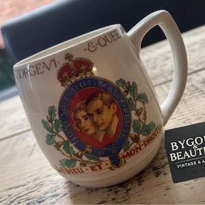 Może przedstawiać: Biały ceramiczny kubek z królewskim herbem z portretami i napisem "GEORGE VI & QUEEN". Kubek ma zakrzywiony uchwyt i jest ozdobiony koroną oraz słowami "DIEU ET MON DROIT".