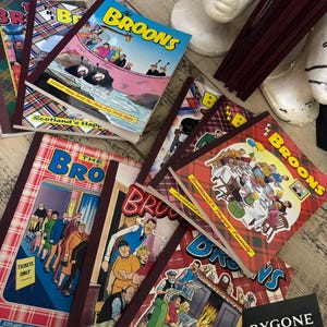 Pode incluir: Uma coleção de revistas em quadrinhos vintage "The Broons", com ilustrações coloridas e o título em amarelo e azul. As capas exibem cenas familiares, emolduradas por um padrão xadrez e lombadas bordô. Dispostas sobre uma superfície de madeira.