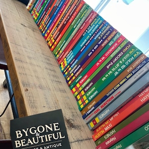 Può includere: Una collezione di manuali di riparazione auto Haynes con dorsi colorati, disposti su una superficie di legno. I libri sono impilati ad angolo, mostrando vari modelli di auto. Un cartello nero con testo bianco recita "BYGONE & BEAUTIFUL VINTAGE & ANTIQUE."