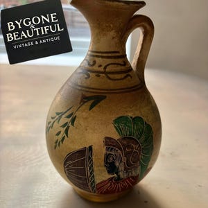 Puede incluir: Jarra de cerámica antigua pintada a mano, con asa y pico. La jarra presenta un color base beige con detalles pintados en marrón, verde y rojo, incluyendo una figura de guerrero y follaje. La jarra tiene el texto "BYGONE & BEAUTIFUL VINTAGE & ANTIQUE".
