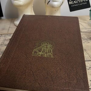 May include: A brown, textured book with a gold embossed design in the center. The book has a rectangular shape and a slightly worn appearance. The cover features a detailed illustration. The book is vintage and antique.