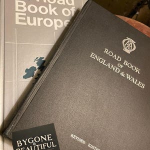 May include: Two vintage road books: "Road Book of Europe" and "Road Book of England & Wales." The books are in gray and black, with white lettering. A small card reads "Bygone & Beautiful Vintage & Antique."