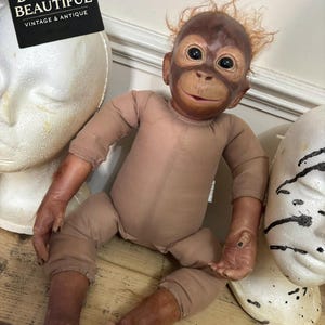 May include: A realistic orangutan doll with a brown vinyl face, hands, and feet, featuring sparse reddish-brown hair and a tan fabric body. The doll has dark eyes and a smiling expression. A black sign with white text reads BYGONE & BEAUTIFUL VINTAGE & ANTIQUE.