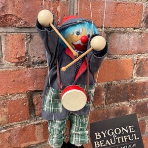 Puede incluir: Una marioneta de payaso vintage con una nariz roja, cabello azul y un atuendo rojo y gris. La marioneta sostiene baquetas y un pequeño tambor. Los pantalones del payaso son de cuadros verdes y tiene zapatos negros. La marioneta está suspendida por cuerdas.
