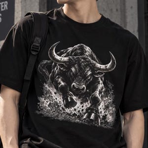 May include: A black short-sleeved t-shirt featuring a detailed graphic print of a charging bull. The bull is rendered in shades of black, white, and grey, depicted in a dynamic forward-facing pose with large horns and hooves kicking up dust.
