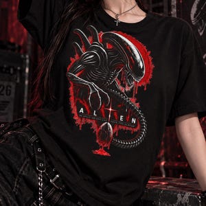 Könnte beinhalten: Ein schwarzes T-Shirt mit einem Grafikdruck der Alien-Kreatur in Schwarz und Weiß vor einem roten kreisförmigen Hintergrund. Unter der Kreatur steht der Text ALIEN und IN SPACE NO ONE CAN HEAR YOU SCREAM, mit einem kleinen, rot tropfenden Ei-Symbol.