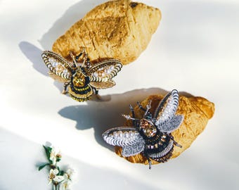 May include: Two decorative bee brooches with intricate detailing. One bee features gold, black, and yellow accents with pearl embellishments. The other bee has silver, black, and white accents with pearl embellishments. Both are displayed on a golden-brown croissant.