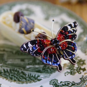 Pode incluir: Broche de borboleta detalhado com lantejoulas e contas pretas, vermelhas e azuis. As asas têm um design intrincado. O broche está sobre um prato branco com um padrão floral verde.