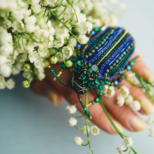 Op de afbeelding: Een geborduurde keverbroche in blauwe en groene tinten, versierd met kleine kralen en een centrale edelsteen. De broche is geplaatst tegen een achtergrond van witte lelietjes-van-dalen, wat een contrast van texturen en kleuren creëert.