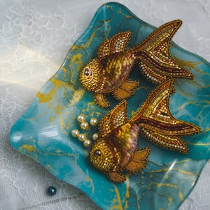 Puede incluir: Dos broches de pez dorado con cuentas doradas, marrones y amarillas descansan sobre un plato turquesa y dorado veteado. Pequeñas perlas están esparcidas alrededor de los peces, creando una exhibición decorativa.