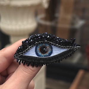 May include: A handcrafted eye-shaped brooch. The iris is a detailed blue and brown, set in white. The eyelid and surrounding area are black, with a beaded texture. The brooch is held in a hand.