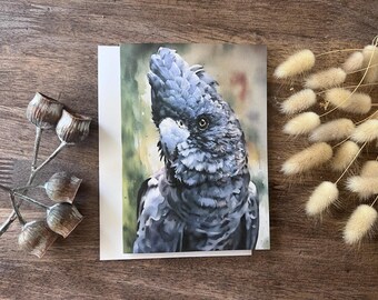 Op de afbeelding: Wenskaart met een aquarel schilderij van een zwarte kaketoe met blauwe en grijze veren. De vogel staat tegen een zachte groene en beige achtergrond. De kaart ligt op een houten oppervlak met gedroogde bloemen en zaaddozen.