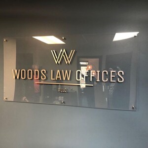 May include: A clear glass sign with gold lettering that reads "WOODS LAW OFFICES PLLC". The sign has a gold "W" logo at the top and is mounted on a gray wall with silver hardware.