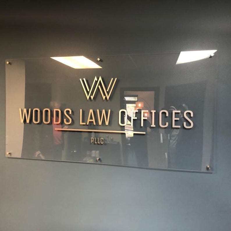 May include: A clear glass sign with gold lettering that reads "WOODS LAW OFFICES PLLC". The sign has a gold "W" logo at the top and is mounted on a gray wall with silver hardware.