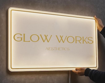 May include: Illuminated rectangular sign with the text "GLOW WORKS" in gold lettering, with "AESTHETICS" below. The sign has a white face and a gold border, held by two hands. The sign is plugged into a wall.
