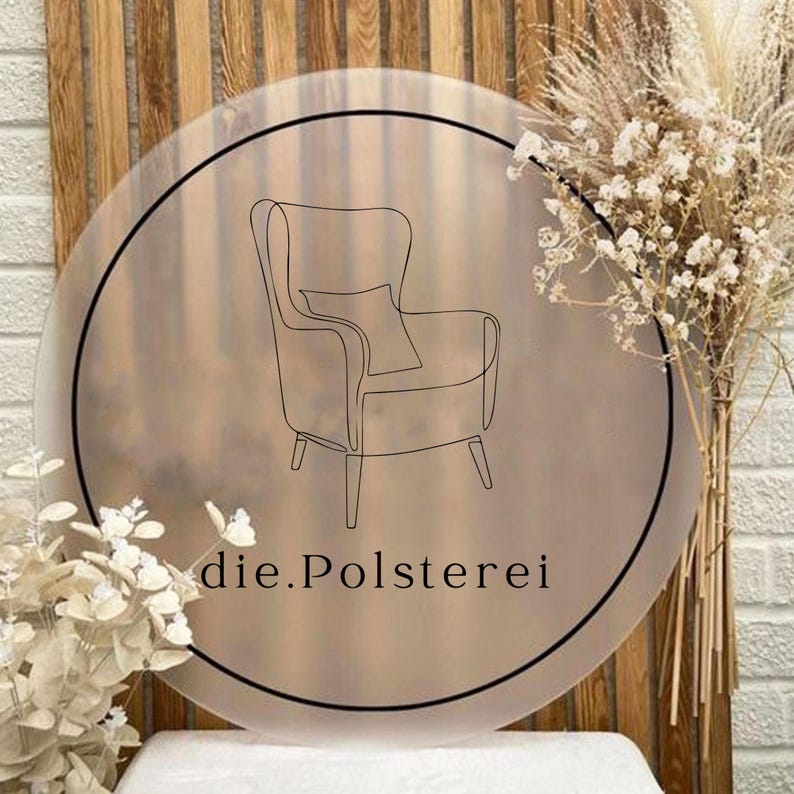 May include: A round sign with a black border features a line drawing of an armchair with a pillow. The text "die.Polsterei" is below the armchair. The sign is set against a backdrop of wood and a white brick wall, with dried flowers.