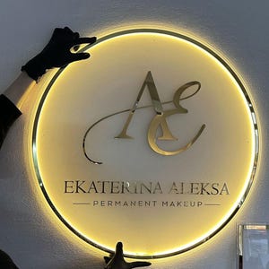 Può includere: Un'insegna rotonda e illuminata con un bordo dorato e un centro color crema. L'insegna presenta le iniziali dorate "AE" e il testo "EKATERINA ALEKSA - PERMANENT MAKEUP".
