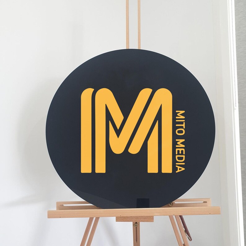 May include: A round black sign with a large yellow "M" logo and the words "MITO MEDIA" in yellow. The sign is displayed on a wooden easel against a white background.