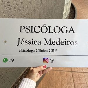 May include: A rectangular white sign with black text. The sign reads "PSIC&Oacute;LOGA J&eacute;ssica Medeiros Psic&oacute;loga Cl&iacute;nica CRP". Below the text are social media icons and a phone number. The sign is held by a person.