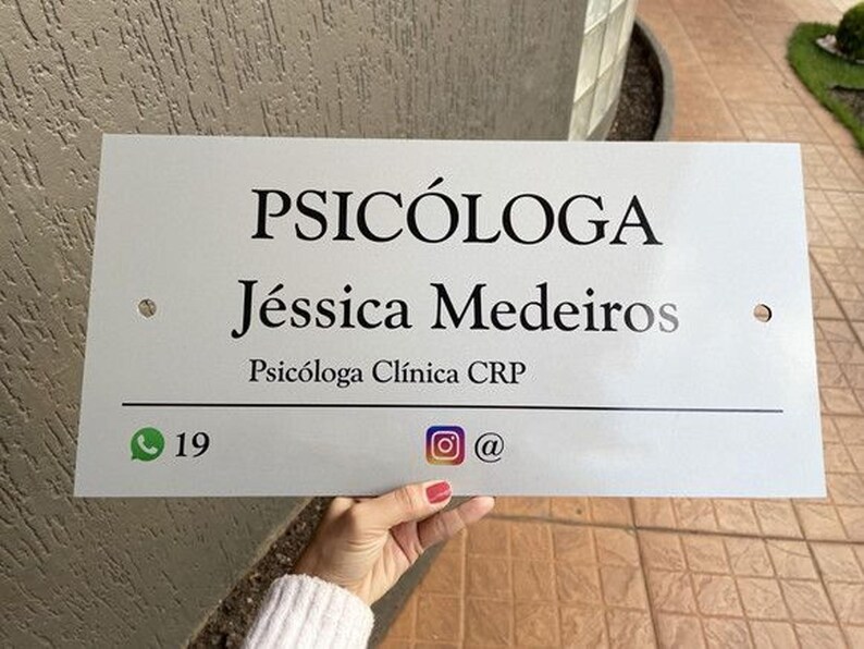 May include: A rectangular white sign with black text. The sign reads "PSIC&Oacute;LOGA J&eacute;ssica Medeiros Psic&oacute;loga Cl&iacute;nica CRP". Below the text are social media icons and a phone number. The sign is held by a person.