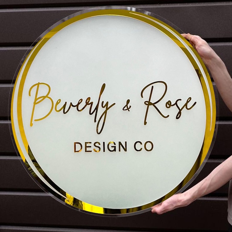 Może przedstawiać: Okrągły, biały znak z podw&oacute;jną złotą obw&oacute;dką. Na znaku widnieje napis "Beverly & Rose DESIGN CO" złotymi literami.
