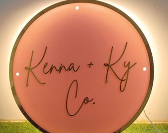 May include: A round, illuminated sign with a coral-colored center and a dark brown border. The sign features the text "Kenna + Ky Co." in a cursive font. The sign is lit from behind, creating a warm glow.