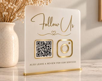 May include: A rectangular white acrylic sign with a gold metal base featuring the gold text Follow Us, a small heart icon, a black and white QR code, a gold Instagram logo, and the text ALSO LEAVE A REVIEW FOR OUR SERVICES in gold lettering.