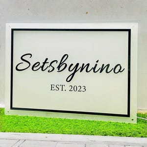 May include: A rectangular sign with the text "Setsbynino" in a cursive font, with "EST. 2023" below it. The sign has a black border and is set against a white background. The sign is placed on a bed of green moss.