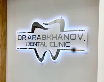 May include: A backlit sign for a dental clinic. The sign features a stylized tooth design above a rectangular panel with the text "DR ARABKHANOV. DENTAL CLINIC" in a modern font. The sign is mounted on a white wall.