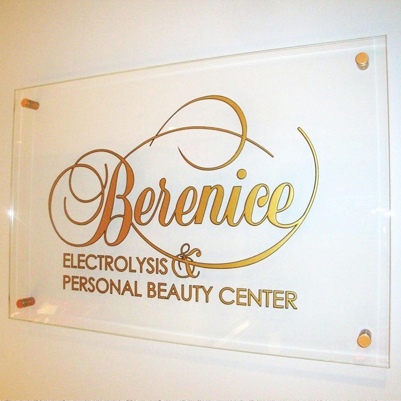 May include: A clear acrylic sign with gold lettering that reads "Berenice Electrolysis & Personal Beauty Center." The sign is mounted on a white wall with four gold-colored standoffs.