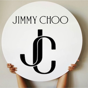 May include: A white, round sign featuring the black text "JIMMY CHOO" above a stylized "JC" logo. The logo is a black, interlocking design of the letters J and C. The sign is held up by two hands.